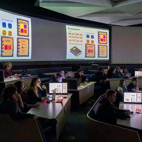A tiered presentation room with rows of desks and computers, where people are watching slides projected on two large screens. The slides display UI components and text.