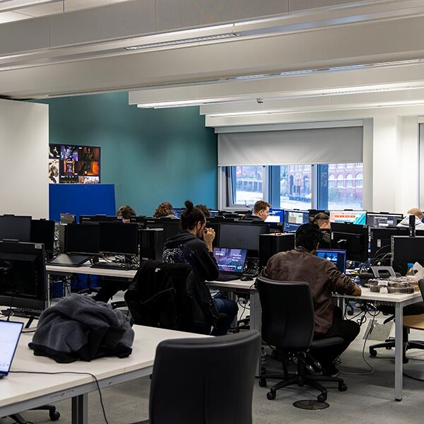 A well-lit office space with rows of computer workstations, where several people are seated and working. The room features white tables, black chairs, and a blue accent wall. A laptop is visible in the foreground.