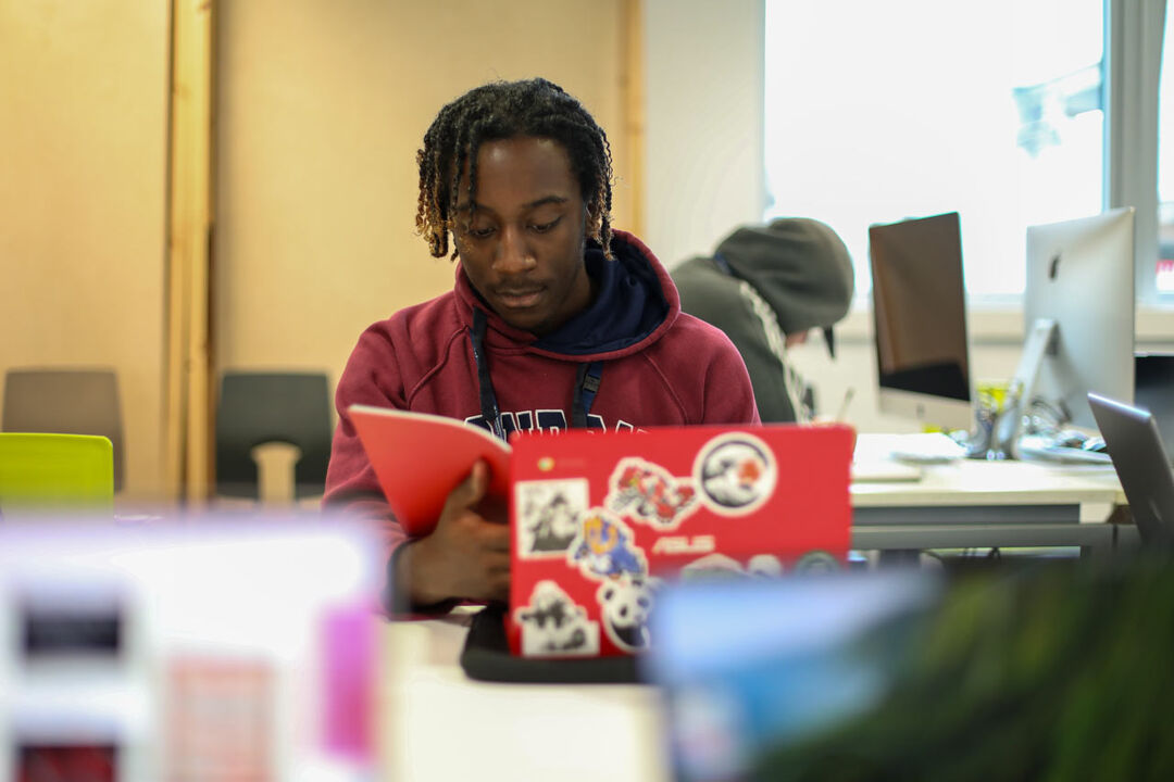 A young man with dark brown hair in twists with lighter ends, who appears to be black sits behind a laptop looking at a book thoughtfully. He is probably studying or working. His laptop is decorated with lots of stickers which are out of focus.