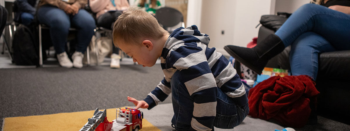 A young boy wearing a blue and white striped hoodie and jeans, crouches on a yellow and grey carpet, playing with a red toy fire truck. In the background, adults are seated, partially visible.