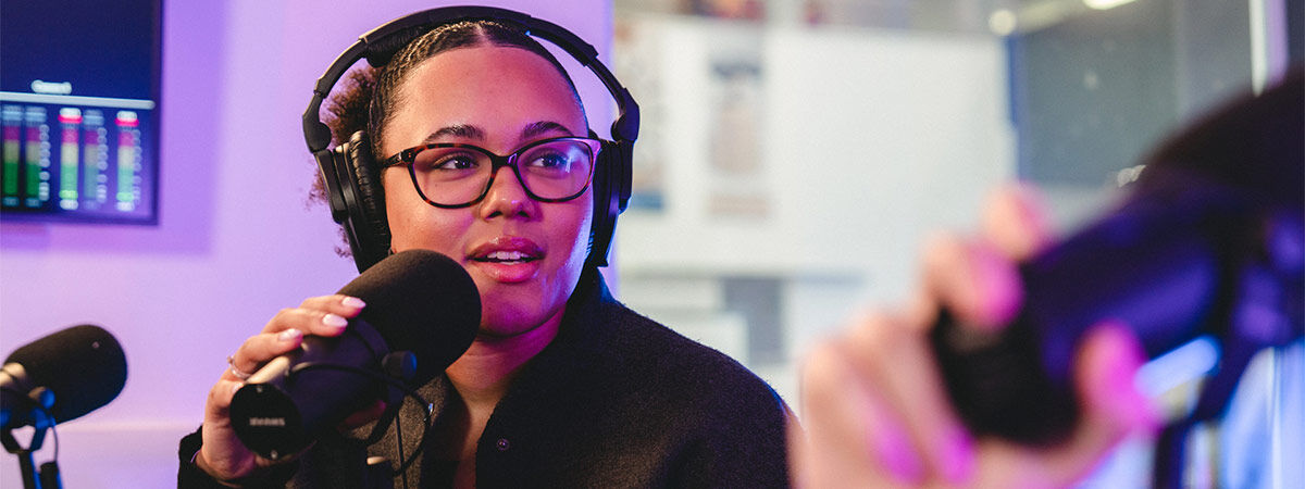 A student is in a radio studio, wearing headphones and speaking into a microphone