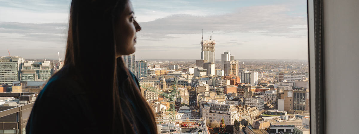 A woman stands by a window in silhouette, looking out over an expansive city skyline. In the distance, several tall buildings are under construction with visible cranes against a clear sky.