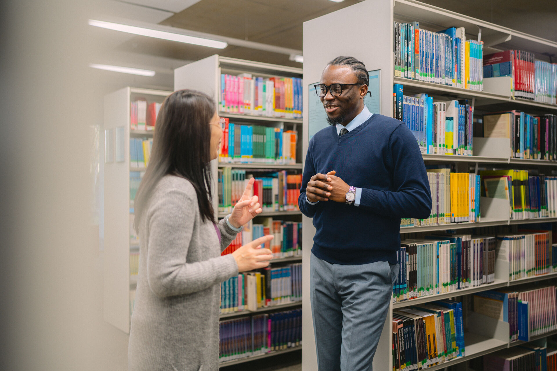 A smartly dressed black man with braids and glasses smiles at a fellow student. They're in the library surrounded by books 