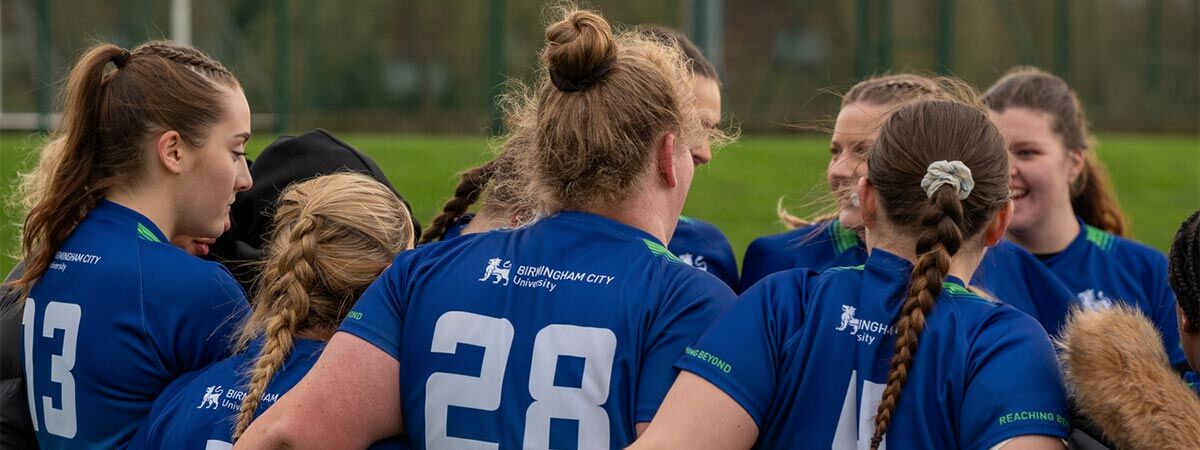 A female rugby team is formed in a huddle on the pitch