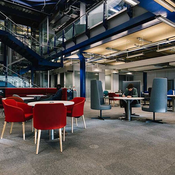 Interior view of a contemporary office space with various seating arrangements, including red chairs around a white table, and tall grey chairs at smaller tables. A staircase is visible in the background.