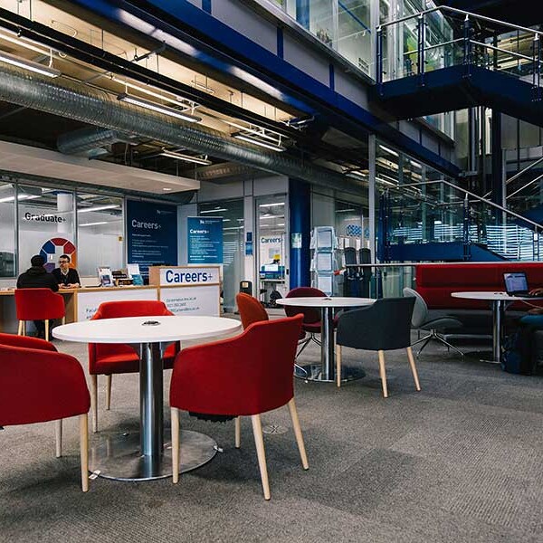 Interior view of a contemporary office space with various seating arrangements, including red chairs around a white table, and people working at tables. A staircase is visible in the background.