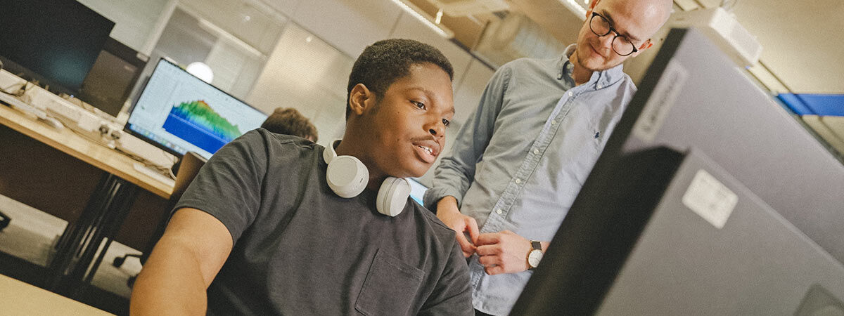 A man with headphones around his neck sits at a computer while another man with glasses stands beside him, both looking at a monitor displaying sound engineering software.