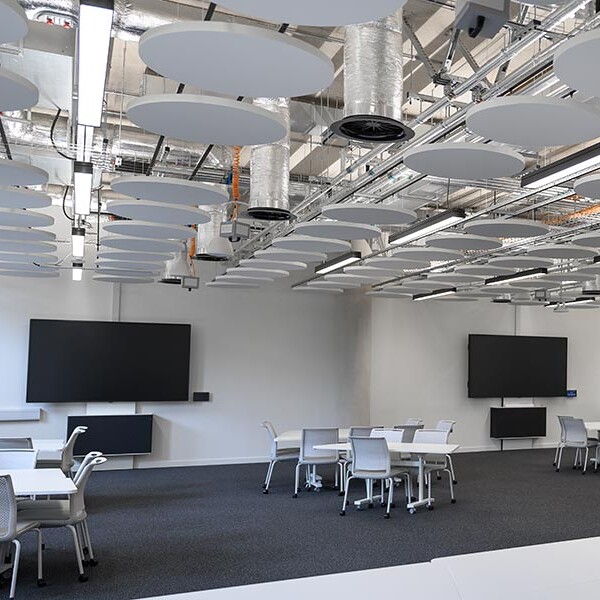 An interior view of a contemporary room with white tables and chairs, featuring two large black screens on the wall and circular acoustic panels on the ceiling.