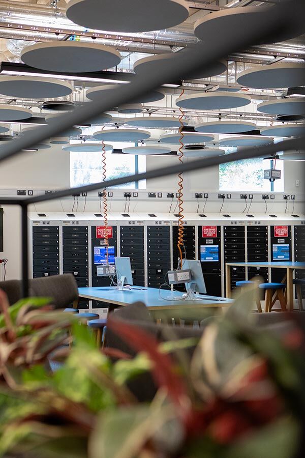 A view of a modern room with computers and storage lockers, through a blurred foreground of plants and railings. The ceiling features circular acoustic panels and visible ductwork.