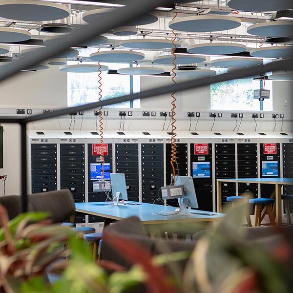 A view of a modern room with computers and storage lockers, through a blurred foreground of plants and railings. The ceiling features circular acoustic panels and visible ductwork.