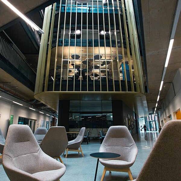 Contemporary lounge space with several light grey upholstered chairs and a small black table, with a view of a multi-level building interior with glass walls.