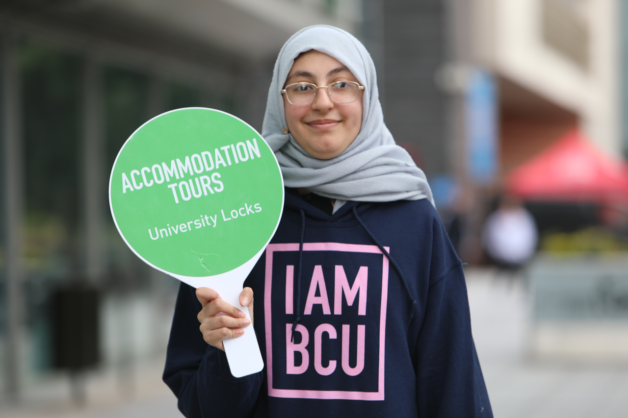 A student ambassador wearing a hijab and glasses holds up a sign which says Accommodation Tours - University Locks