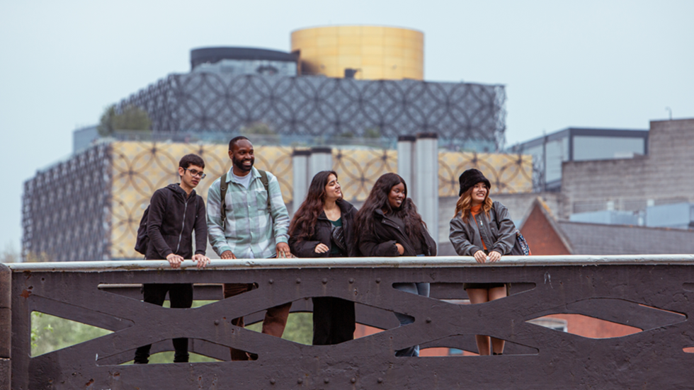 A diverse group of students standing on a canal bridge with the iconic Library of Birmingham in the background. They are smiling and looking towards something out of frame