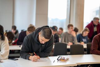 A student who appears to be male with white skin and very short hair writes on a piece of paper with a pencil. He is in a busy classroom and could be taking a test, studying or working. A clear pencil case sits on the table next to him.