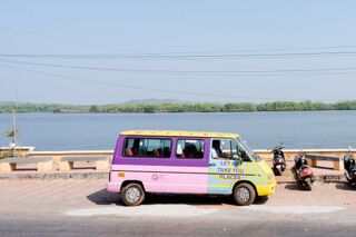 A small van with a pink, purple, yellow, and blue body parked on a paved surface next to a body of water with trees in the background.