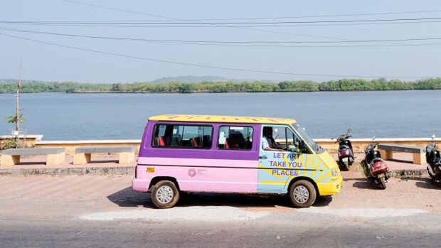 A small van with a pink, purple, yellow, and blue body parked on a paved surface next to a body of water with trees in the background.