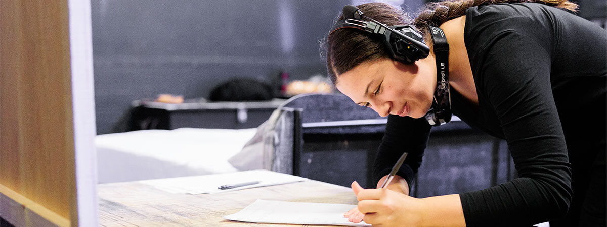 A student is backstage, wearing headphones and writing on a notepad