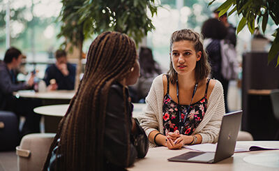 Two students having a discussion in a communal work space with laptop on table.