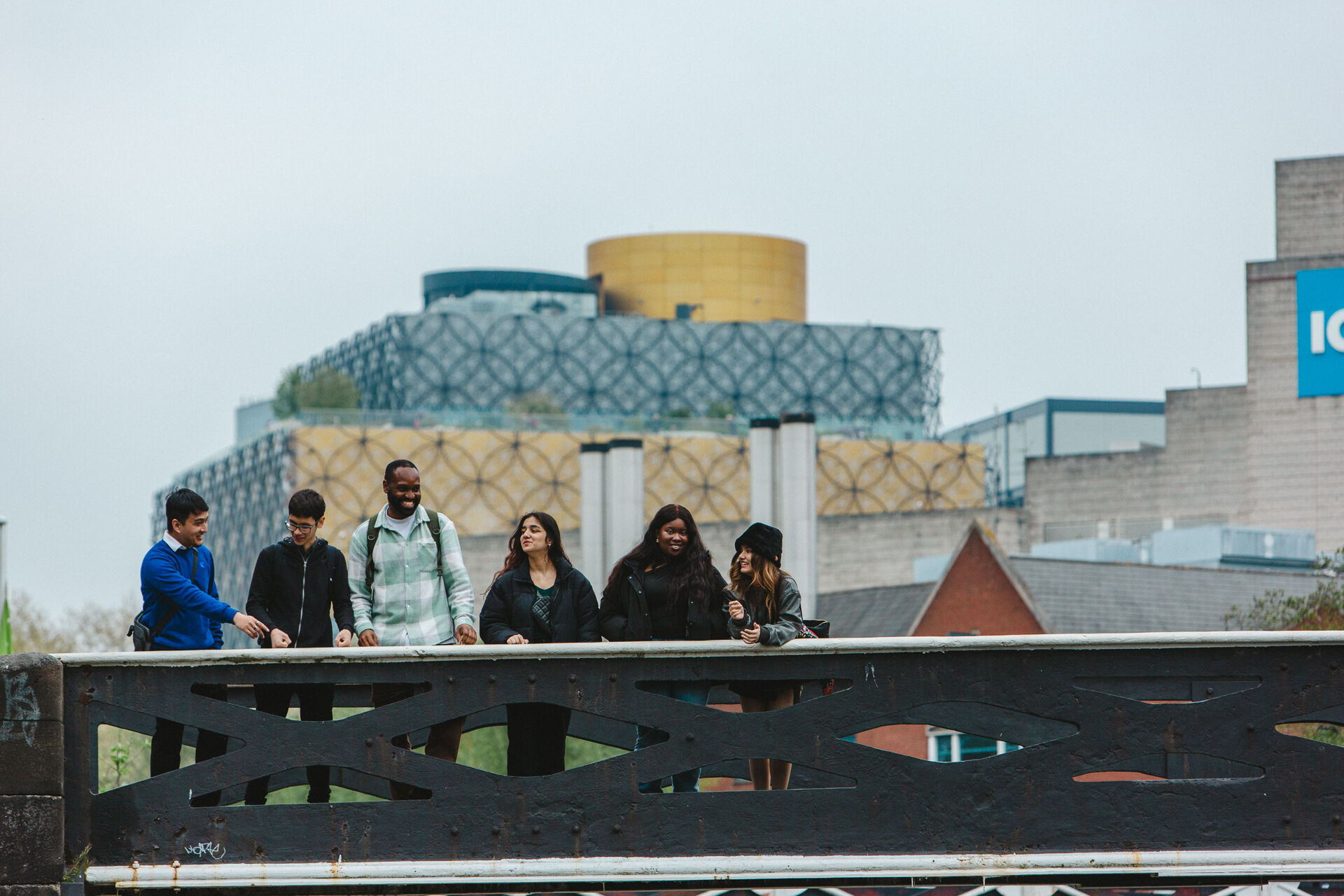 A diverse group of students standing on a canal bridge with the iconic Library of Birmingham in the background. They are smiling and looking towards something out of frame