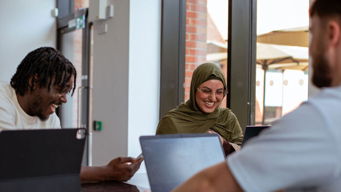 Three students laugh and talk sitting at a desk with laptops