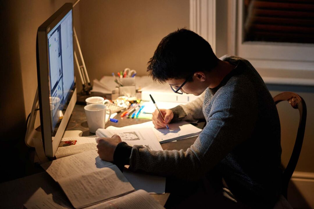 A young person who appears to be male with white skin, wearing glasses and a grey jumper is studying late at night at a cluttered desk. He is writing in a notebook with a pen, surrounded by open books, notes, colourful pens, and two mugs. A computer monitor is on his desk, displaying a webpage, while a desk lamp casts a warm glow over his workspace. The room appears dimly lit, with a window and blinds visible in the background.
