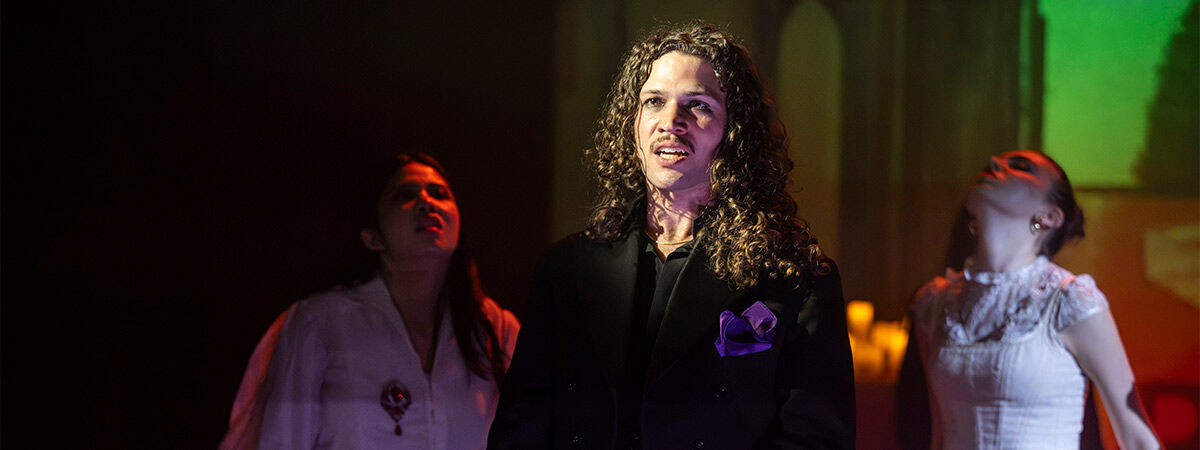 3 BA Acting students performing onstage, with 1 actor dressed in black in the foreground and 2 actors dressed in white in the background