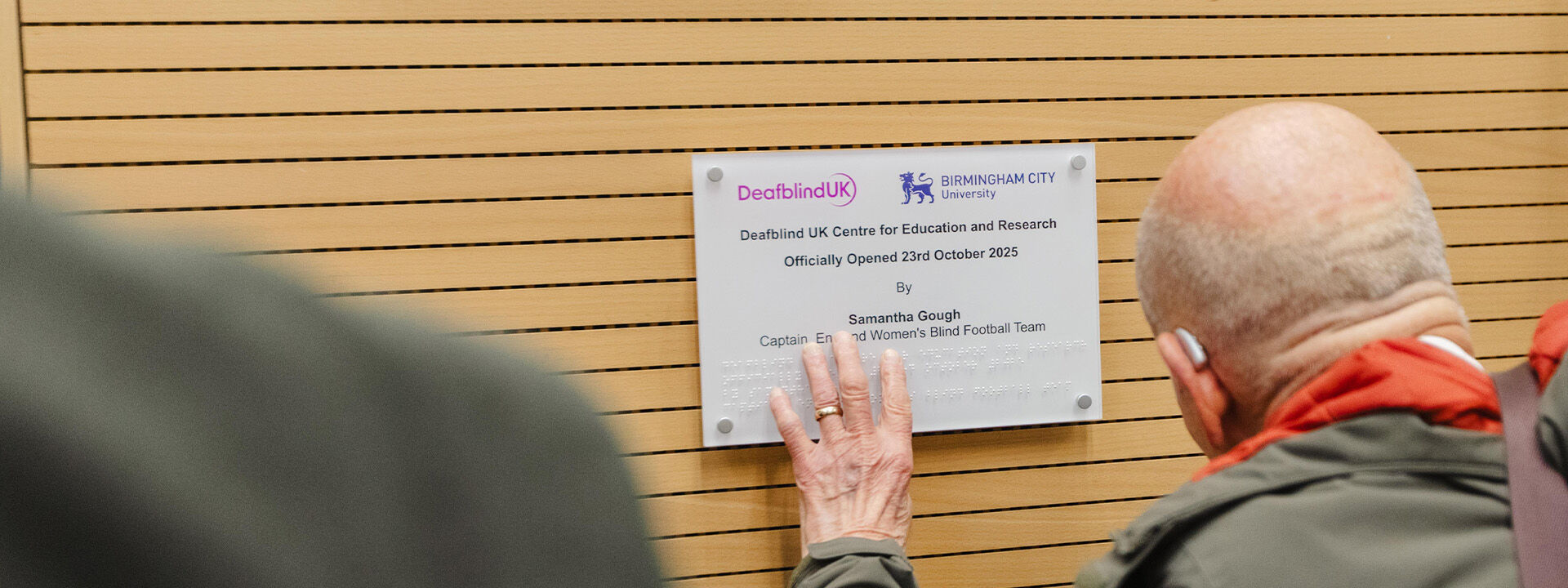 A visually impaired bald man touches brail on a sign reading "Deadblind UK Centre for Education and Research"