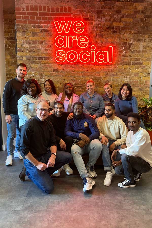 MA Future Media students and lecturer stood and sat in front of neon red sign which reads 'we are social', with a brick background