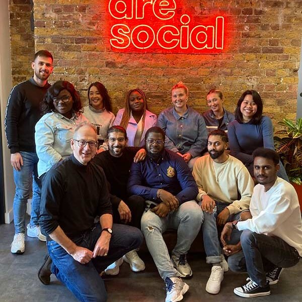 MA Future Media students and lecturer stood and sat in front of neon red sign which reads 'we are social', with a brick background