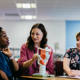 Three women sit at a table in discussion, with one woman pointing to a model of a human larynx and throat.