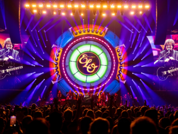 Jeff Lynne's ELO A concert stage with colorful circular lights, ELO logo on a large screen, musicians performing, and an audience watching. Two side screens show Jeff Lynne singing