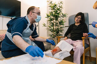 A person in a blue uniform and mask is kneeling to look at the leg of someone sitting in a blue chair. The seated person has a wound on their leg. Another person in blue gloves is holding the seated person's hand. They are inside a room with a sofa and a plant.
